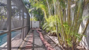 Un espace piscine agrémenté d’un mur en verre, d’un chemin en briques et de diverses plantes tropicales.