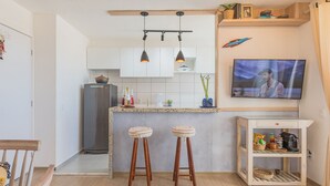 A compact kitchen with a central island, two bar stools, a TV on the wall, and a small shelf with kitchen items. - Mirante #605 - Space and Comfort by Carpediem (Fortaleza)