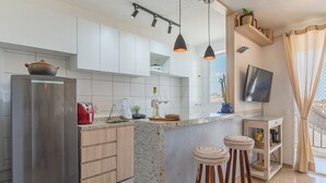 A modern kitchen with a stainless steel refrigerator, wooden cabinets, a granite countertop, pendant lights, a TV mounted on the wall, and a window with curtains. - Mirante #605 - Space and Comfort by Carpediem (Fortaleza)