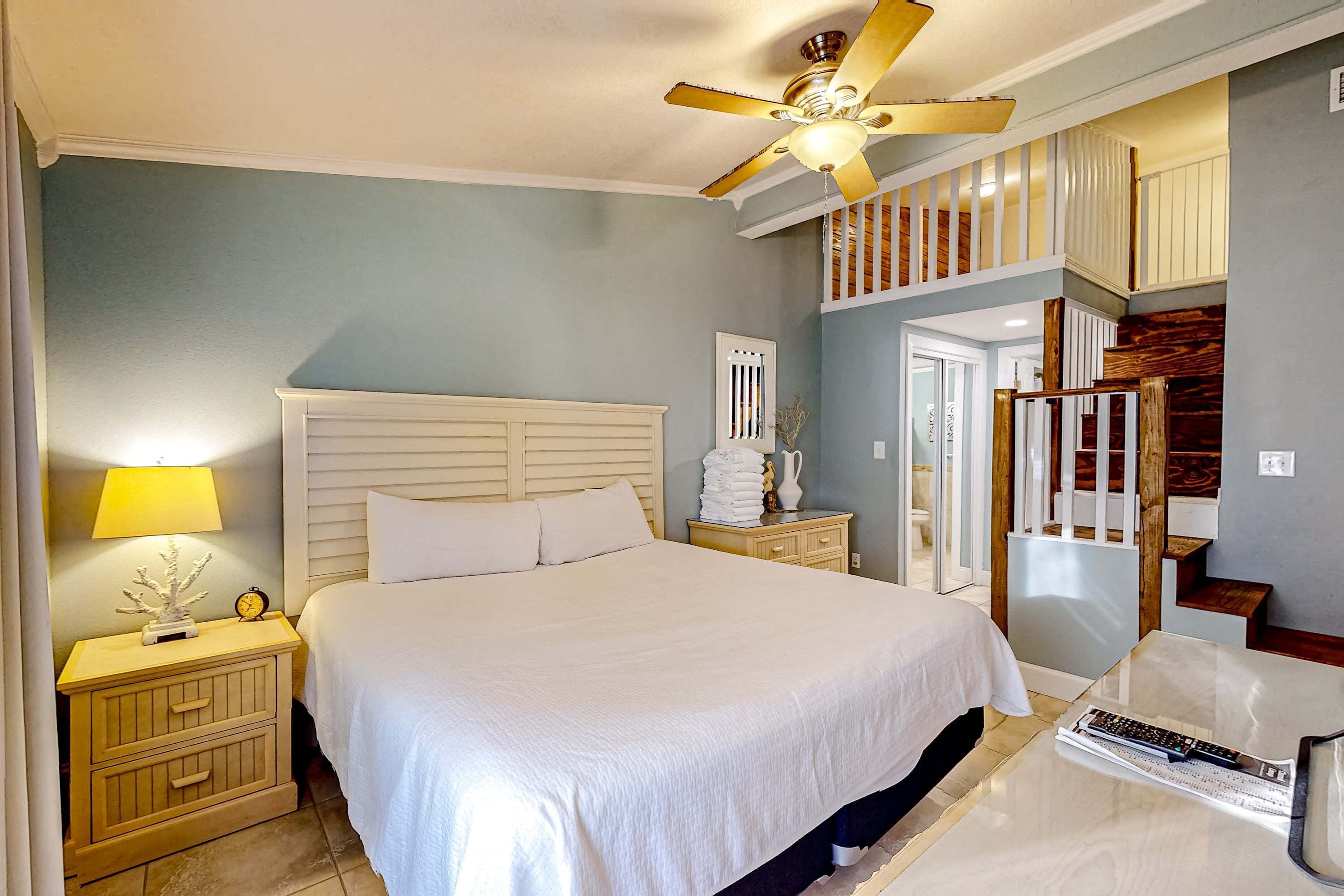 A bedroom with a bed, nightstands, a lamp, a ceiling fan, a staircase, and a door.