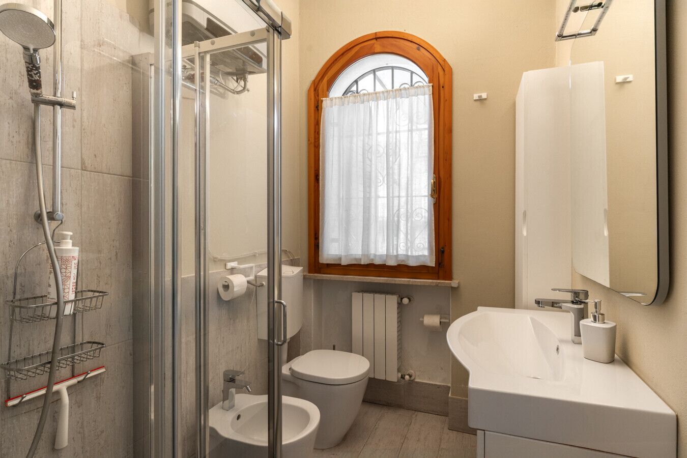 Ein Badezimmer mit Duschkabine aus Glas, einem Bidet, einer Toilette, einem Waschbecken mit Spiegel und einem Fenster mit Holzrahmen.