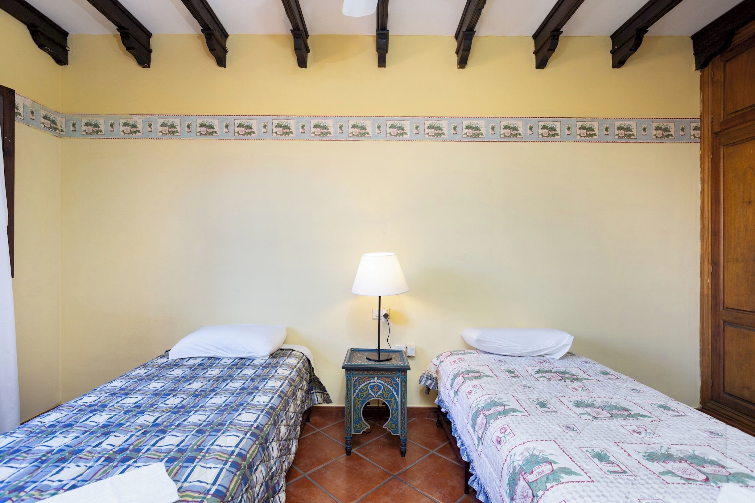 A room with two beds, a small table with a lamp, and a decorative border on the wall.