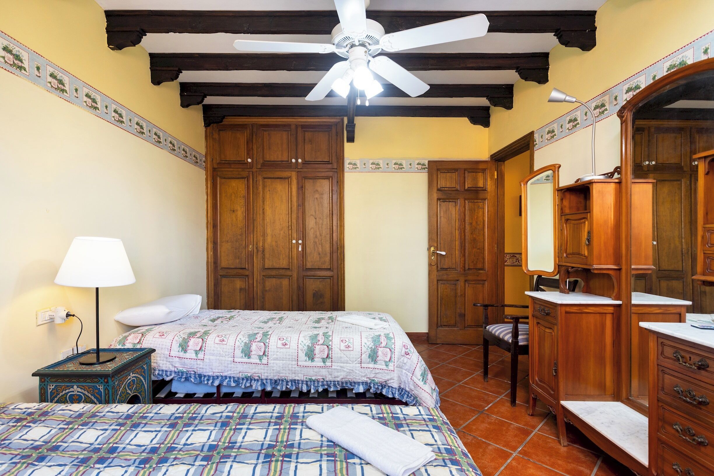 Une chambre à coucher comprenant un lit, une table de chevet, une lampe, une armoire en bois, un miroir et un ventilateur de plafond.