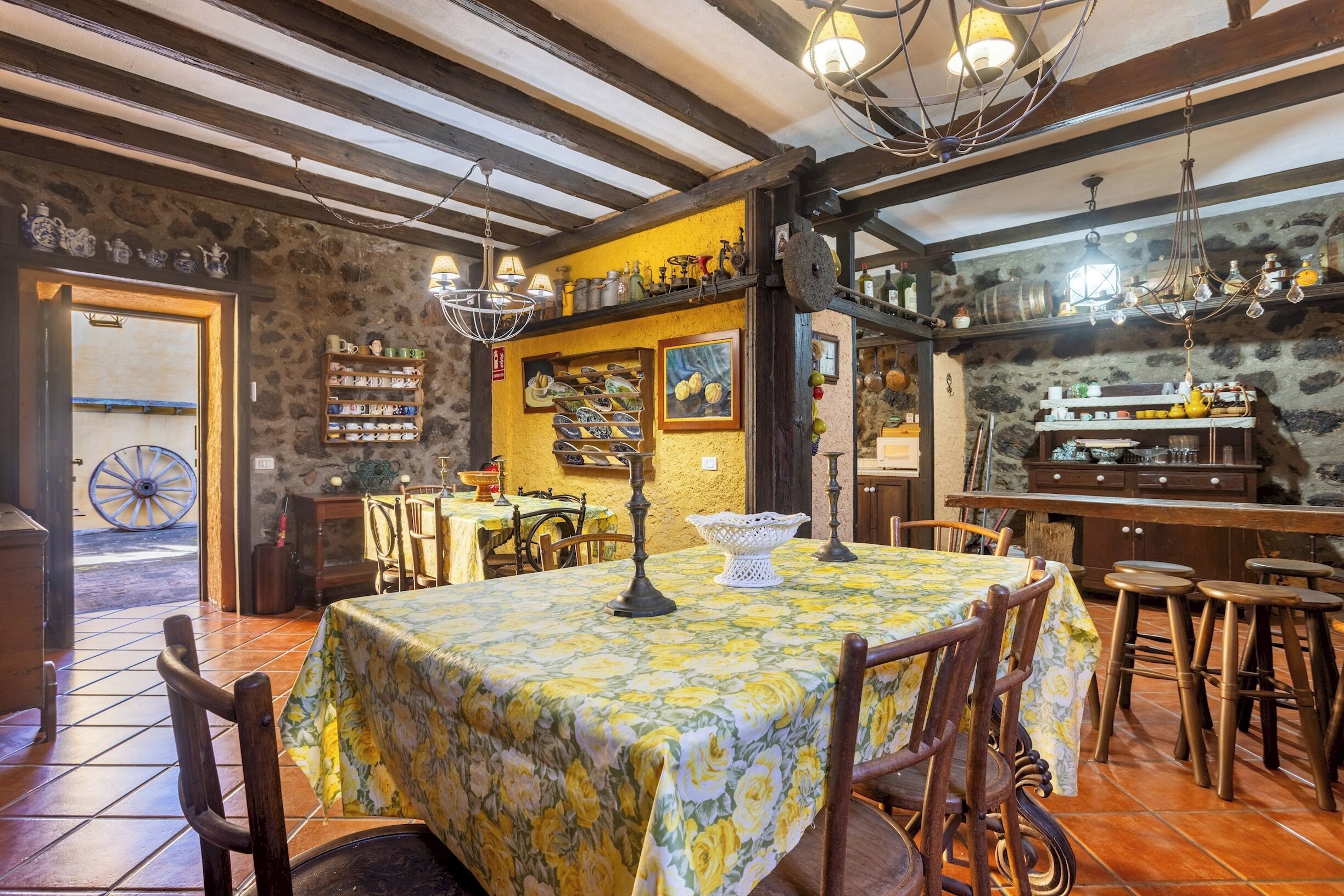 Un comedor rústico con una mesa dispuesta para cuatro personas, rodeada de sillas de madera. Un candelabro cuelga del techo y estanterías exhiben distintos utensilios de cocina.