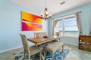 A dining area with a wooden table, chairs, a chandelier, a colorful abstract painting, and a view of the ocean through a window. - Beachfront townhome with ocean views, 2 balconies, W/D, & AC - dog-friendly (Pensacola Beach)