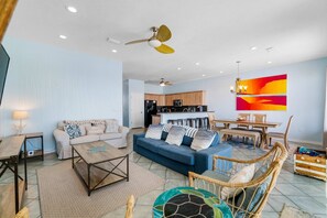 A spacious living room with a blue sofa, a coffee table, and a dining area with a table and chairs. A kitchen is visible in the background. - Beachfront townhome with ocean views, 2 balconies, W/D, & AC - dog-friendly (Pensacola Beach)
