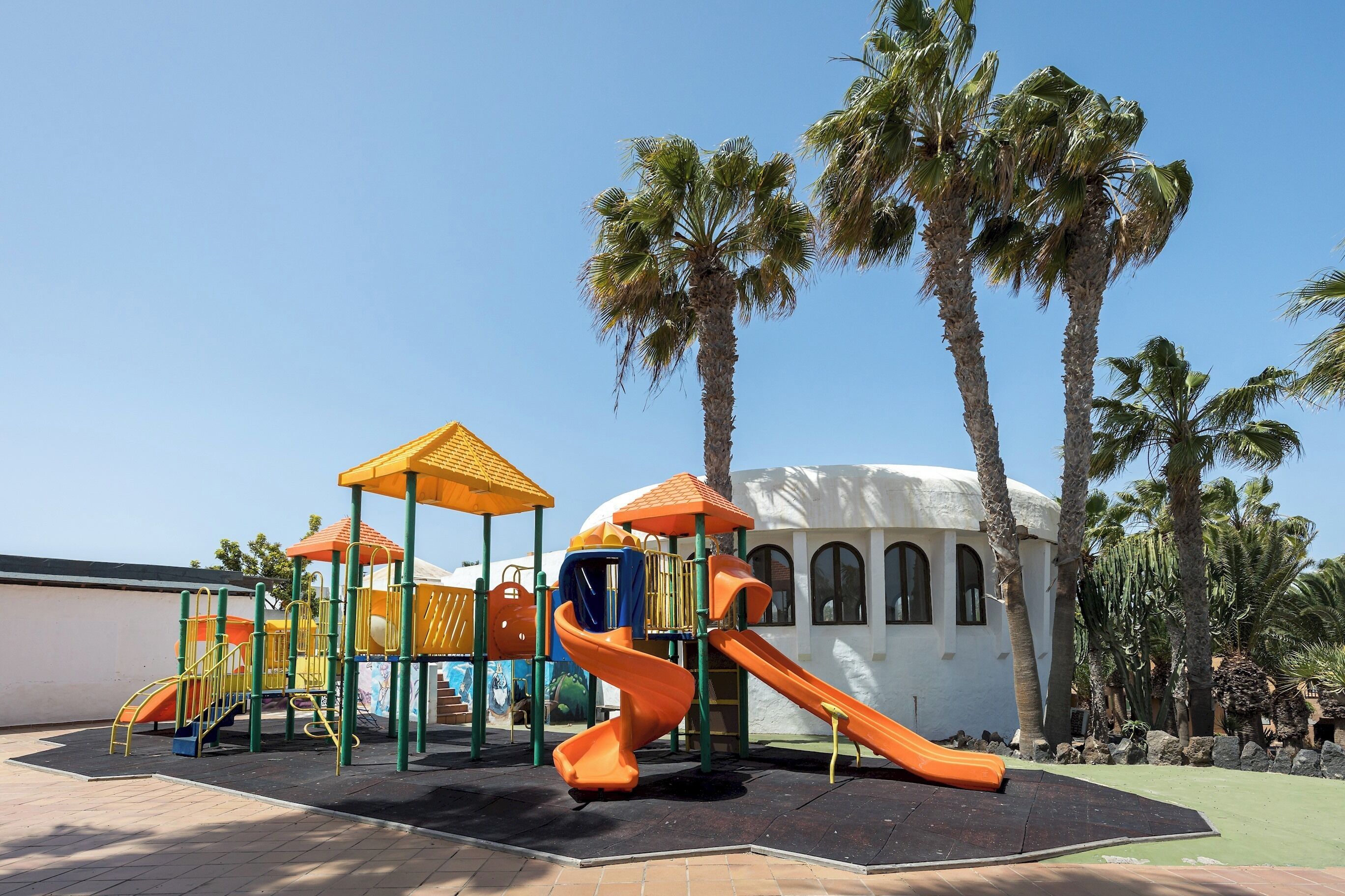 A playground with a slide and climbing structures, surrounded by palm trees and a building with arched windows.