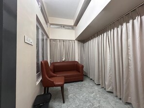A room with a brown sofa, a window with curtains, a light switch, and a trash bin. - NK GUEST HOUSE (Guwahati)