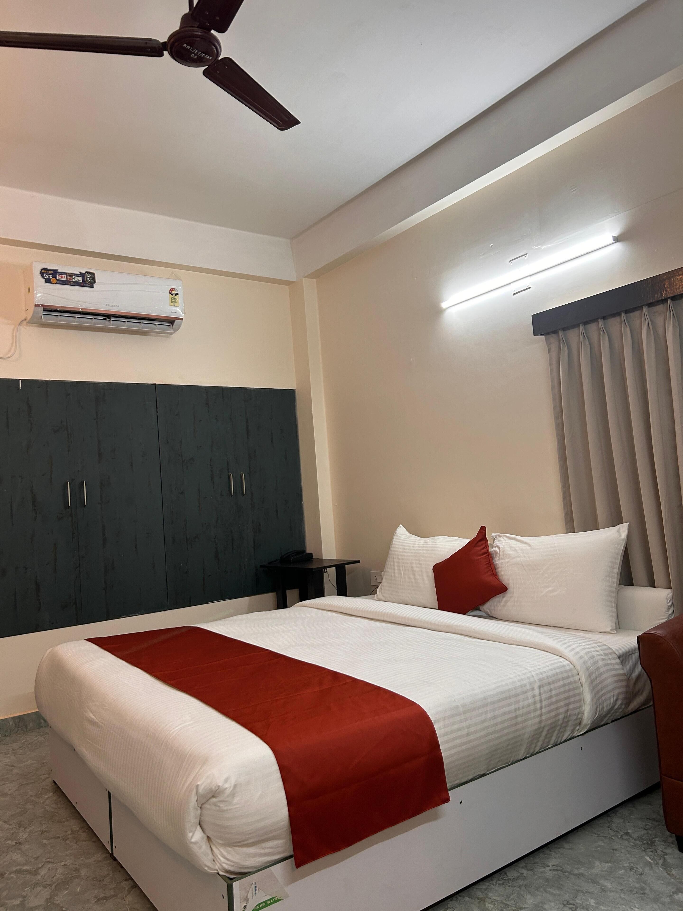A hotel room with a bed, a ceiling fan, an air conditioning unit, a bedside table, and a window with curtains.