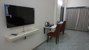 A hotel room with a flat-screen TV mounted on the wall, a small table with a kettle, cups, and glasses, and two brown chairs. - NK GUEST HOUSE (Guwahati)