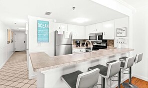 A modern kitchen with a stainless steel refrigerator, a microwave, and a coffee maker. There are bar stools and a tiled floor. - 6th Floor Condo, On-site restaurant & bar, Splash pad w pool (Destin)