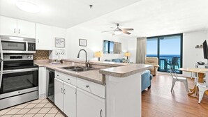 Una cucina moderna con forno a microonde, forno e lavandino integrati. C'è una zona pranzo con tavolo e sedie, e un balcone con vista sull'oceano.