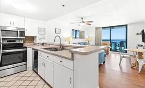 A modern kitchen with a built-in microwave, oven, and sink. There is a dining area with a table and chairs, and a balcony with a view of the ocean. - 6th Floor Condo, On-site restaurant & bar, Splash pad w pool (Destin)