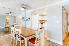 Un coin repas avec une table en bois et des chaises blanches, une cuisine avec des meubles blancs et un couloir avec une porte.