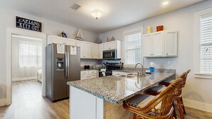 A kitchen with white cabinets, a granite countertop, a stainless steel refrigerator, a microwave, and a sink. There are wicker chairs at the island. - Beautiful home in a gated community with porches & shared pool,hot tub (Port St. Joe)