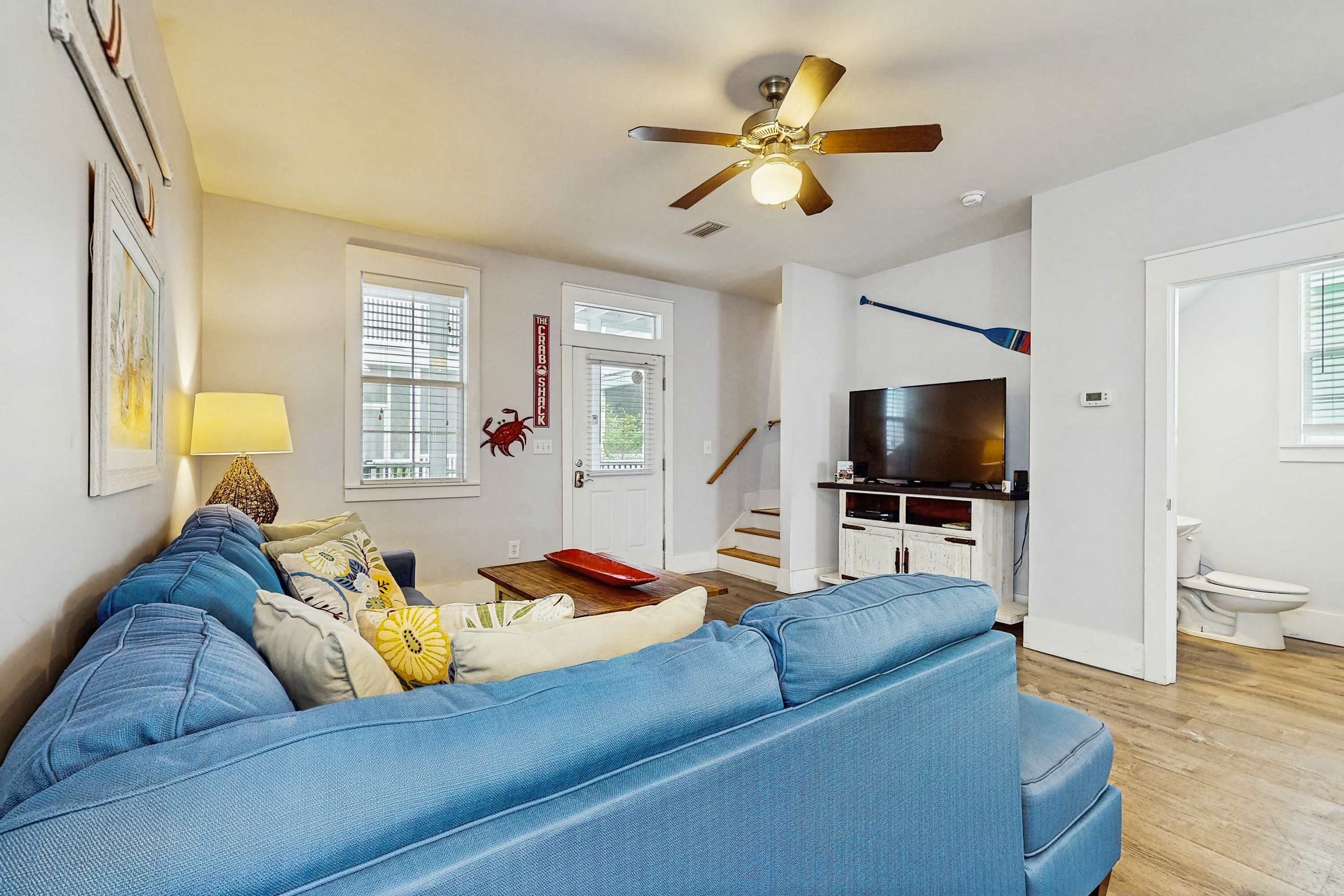 A modern living room with a blue sofa, a ceiling fan, a TV, and a staircase leading to another level.