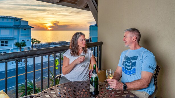 A balcony with a sunset view, a table with a bottle of champagne, and two people seated.