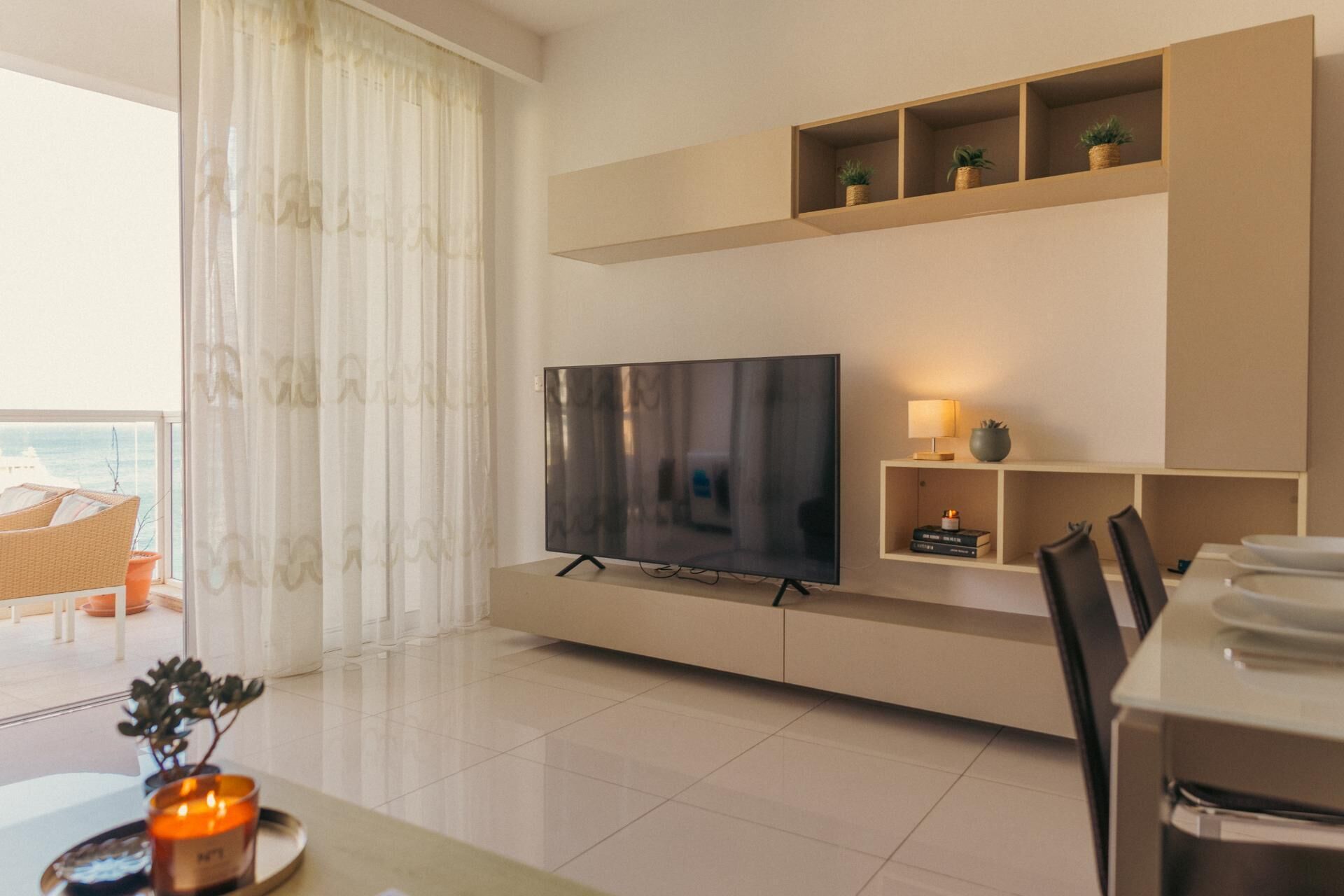 A modern living room with a large flat-screen TV, a dining area with chairs, and a view of the sea through sheer curtains.