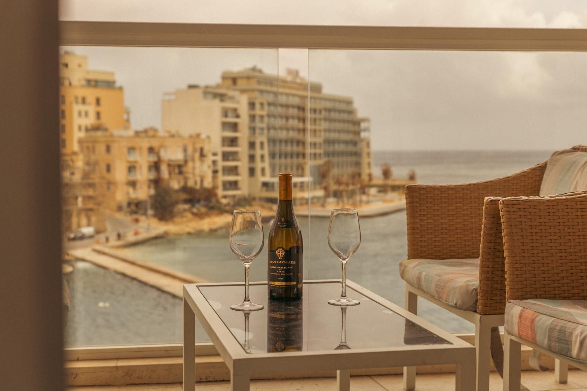A balcony with a table, two wine glasses, and a bottle of wine. The view includes a body of water and buildings in the background.