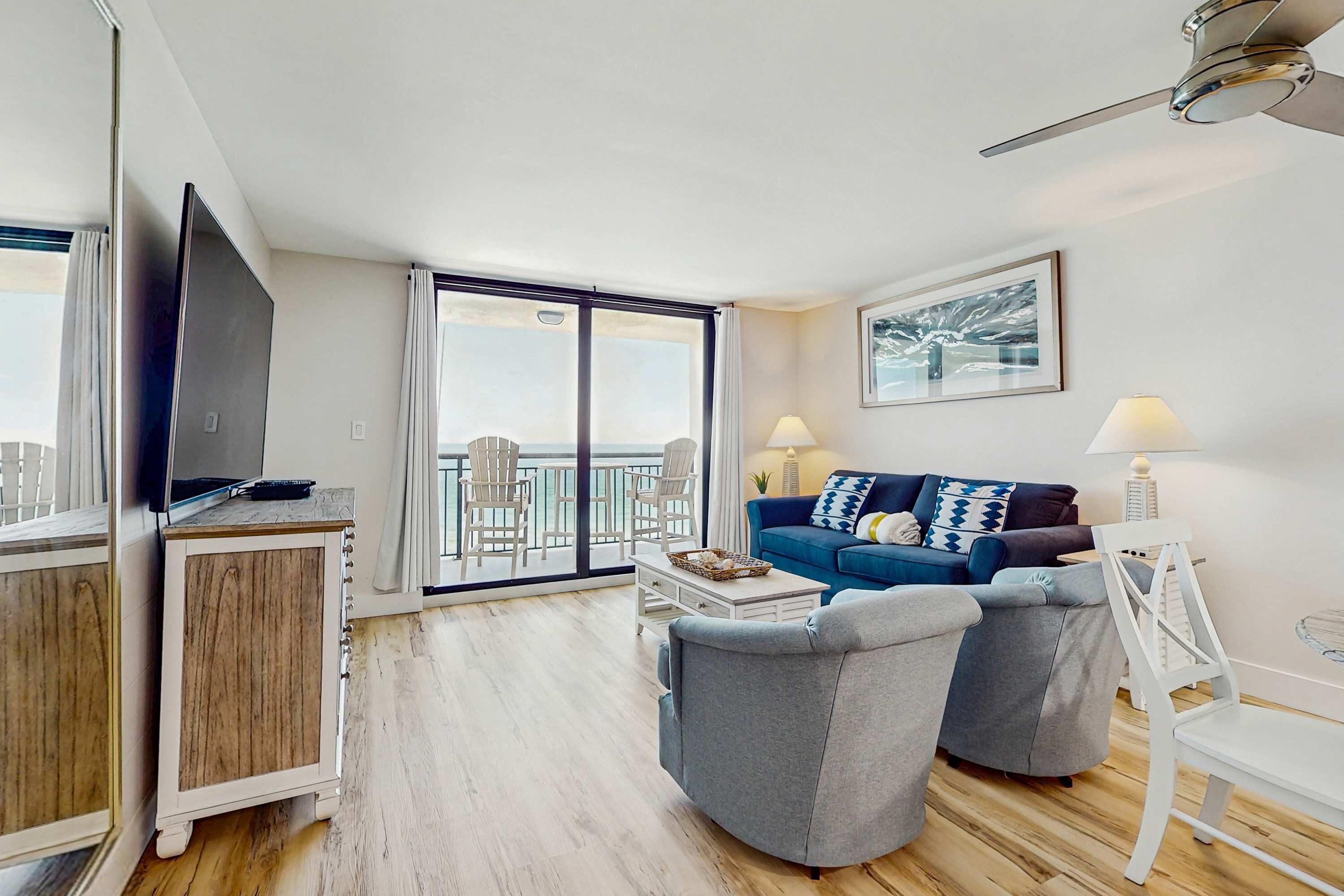 A modern living room with a flat-screen TV, a blue sofa, two grey armchairs, a coffee table, and a balcony with a view.