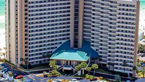 A high-rise hotel with multiple balconies, a central entrance with a green roof, and a parking area with cars. - Spacious oceanfront condo with pool on-site, ocean view, WiFi & central AC (Destin)