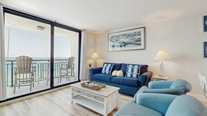 A spacious living room with a balcony, a blue sofa, and a white coffee table. - Spacious oceanfront condo with pool on-site, ocean view, WiFi & central AC (Destin)