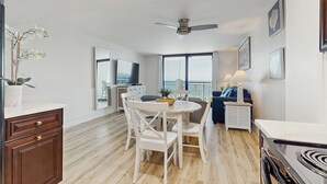 A spacious living area with a dining table, white chairs, a ceiling fan, and a large window offering a view. - Spacious oceanfront condo with pool on-site, ocean view, WiFi & central AC (Destin)