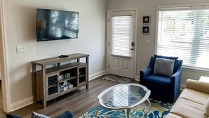 Ett vardagsrum med en platt-TV, ett träinrett TV-bord, ett soffbord i glas, en blå fåtölj och en beige soffa.