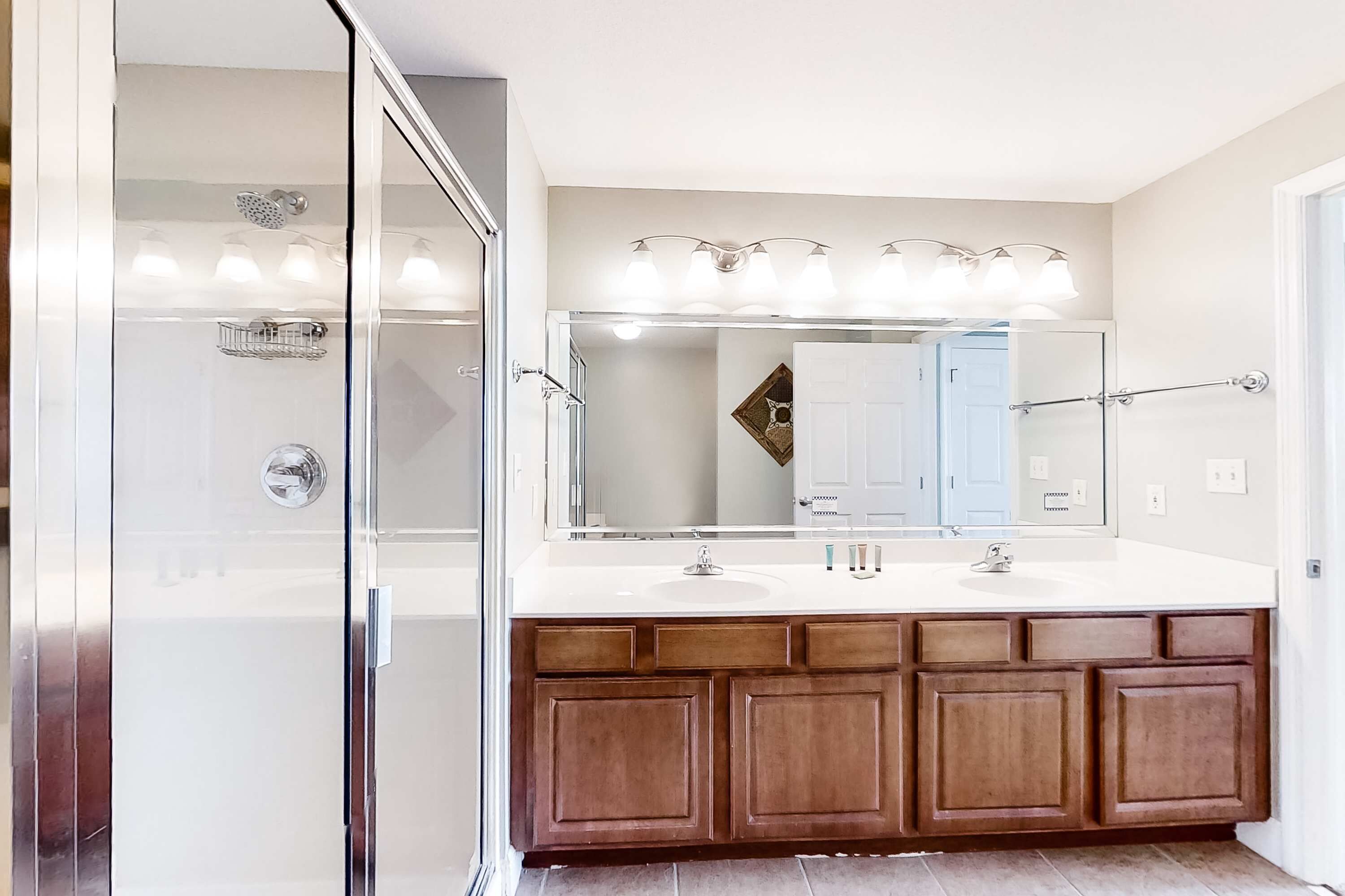 Une salle de bain dotée d’une douche à parois de verre, d’un grand meuble-lavabo avec un miroir et des luminaires, et de meubles de rangement en bois.