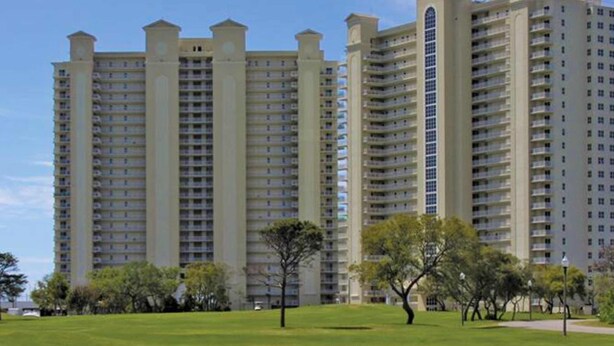 A high-rise residential building with multiple balconies, surrounded by a well-maintained lawn and trees. - Spacious 3 Bedroom Condo with Beach Views and Resort Pool Access (Miramar Beach)