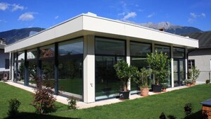 A modern house with a glass conservatory, surrounded by a well-maintained lawn and potted plants. - Hotel Rettenberg (Kolsass)