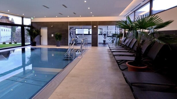 An indoor swimming pool with a clear view of the pool area, a seating area with chairs, and a staircase leading to an upper level. - Hotel Rettenberg (Kolsass)