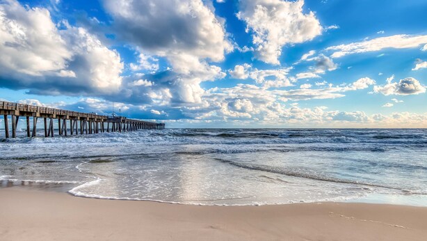 Ein Strand mit einer Holzsteg, der sich ins Meer erstreckt, sanfte Wellen am Ufer und ein teils bewölkter Himmel.