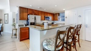 A modern kitchen with wooden cabinets, a central island, and a dining area with chairs. - Corner-condo with wraparound deck, beach access & pool/hot tub (Panama City Beach)