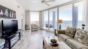 A spacious living room with a flat-screen TV, a sofa with patterned cushions, a coffee table with a decorative bowl, and large windows offering a view of the sea. - Corner-condo with wraparound deck, beach access & pool/hot tub (Panama City Beach)