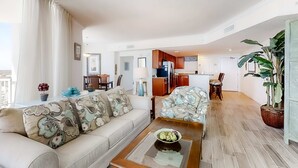 A modern living room with a sofa, coffee table, and a dining area with chairs. There is a kitchen in the background with cabinets and appliances. - Corner-condo with wraparound deck, beach access & pool/hot tub (Panama City Beach)