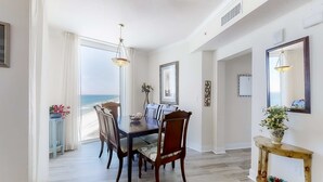 A dining area with a view of the beach, a wooden dining table, chairs, and a console table with a vase. - Corner-condo with wraparound deck, beach access & pool/hot tub (Panama City Beach)