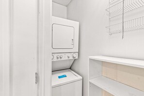 A laundry room with a front-loading washer and dryer, a metal drying rack, and a shelf unit. - Beachview Condo with full kitchen, shared pool and beach access (Panama City Beach)