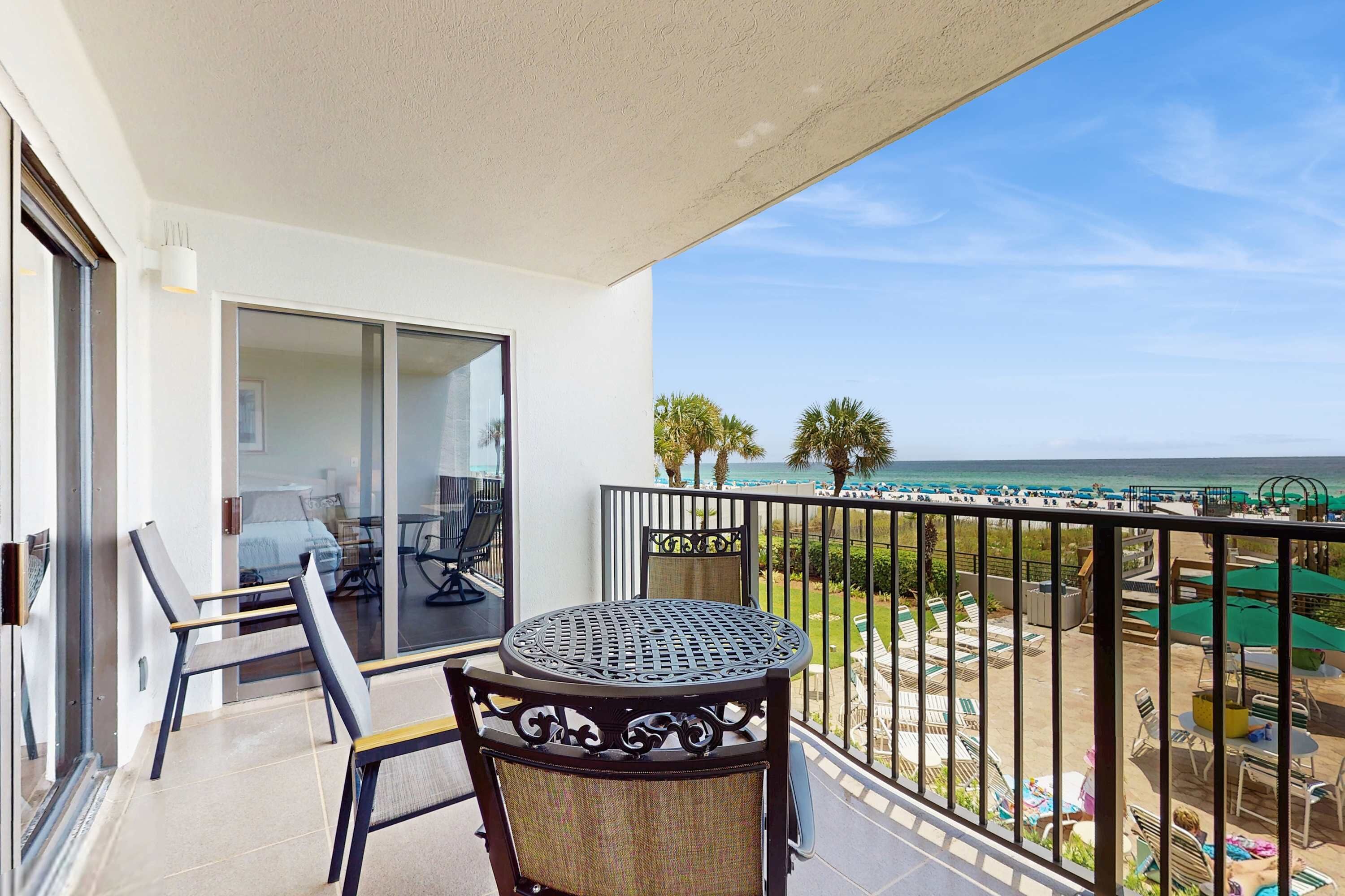 Varanda com mesa e cadeiras, com vista para uma praia com guarda-sóis e espreguiçadeiras.