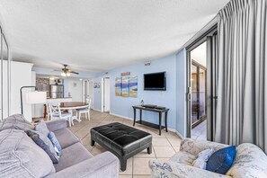 A living room with a grey sofa, a black ottoman, and a patterned armchair. There is a dining table with chairs, a kitchen area, and a TV mounted on the wall. - Ocean-view condo at SunDestin with pool, hot tub, sauna, and walkable locale (Destin)