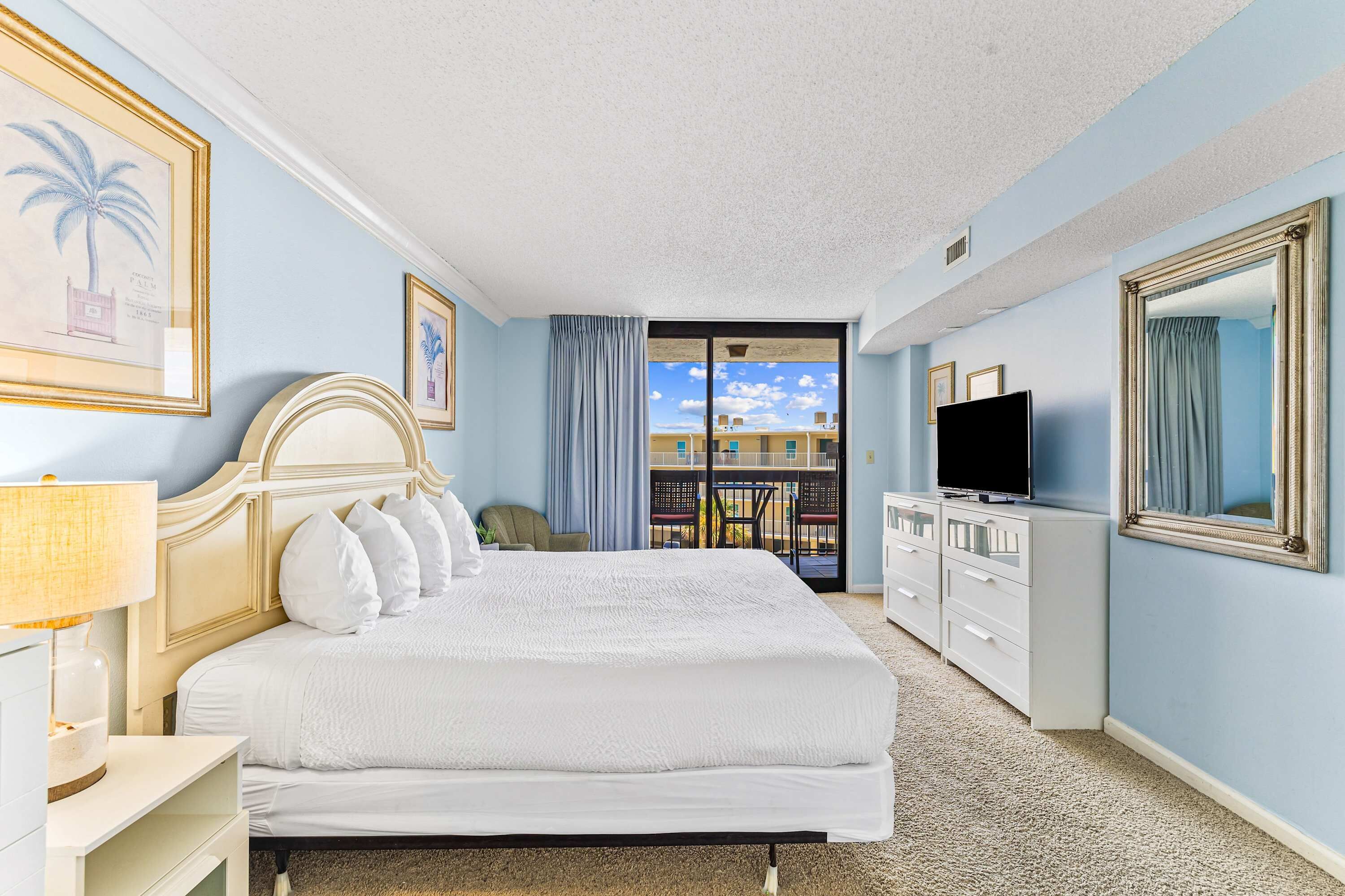A hotel room with a large bed, a television, a dresser, and a balcony with a view of buildings.