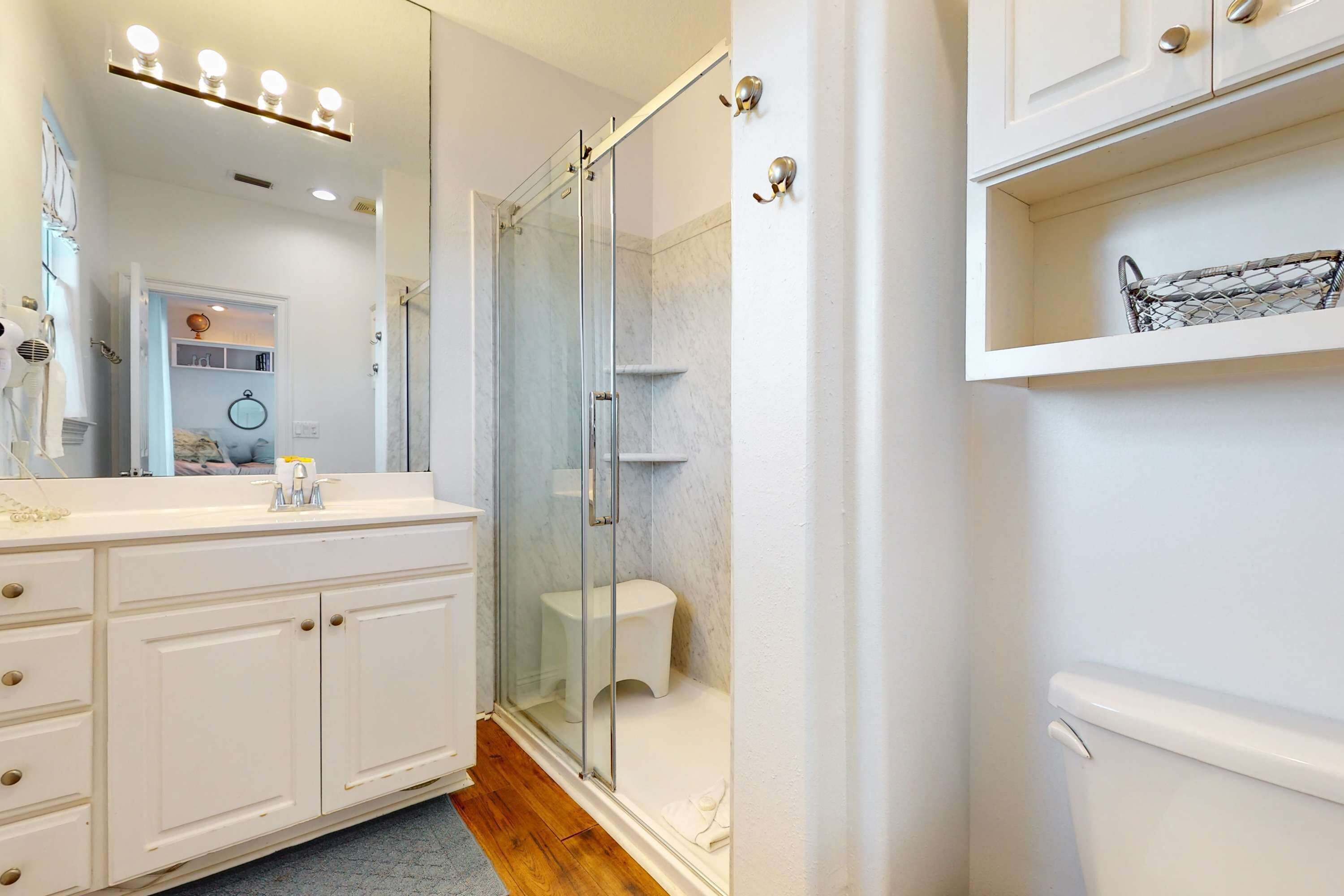 A bathroom with a glass shower enclosure, a white vanity with drawers, a mirror with lights, and a shelf with a basket.