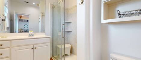 A bathroom with a glass shower enclosure, a white vanity with drawers, a mirror with lights, and a shelf with a basket.