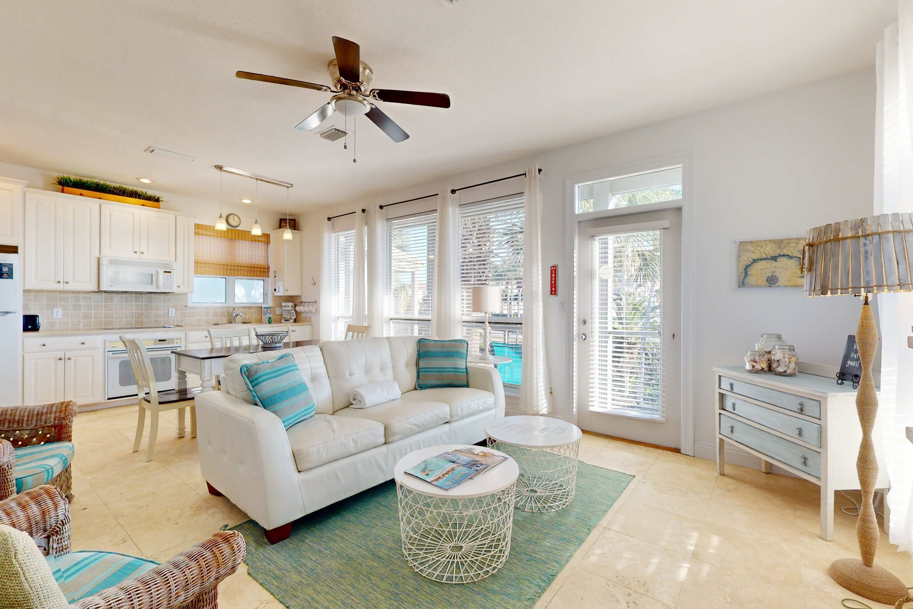A spacious living area with a white sofa, two round side tables, a ceiling fan, and a view of the kitchen and pool through large windows.