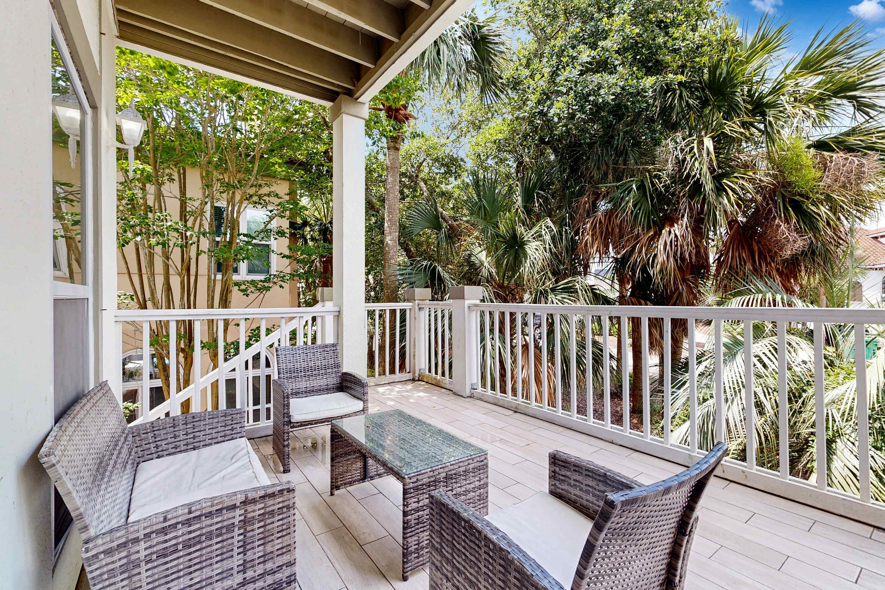 A covered outdoor seating area with wicker furniture and a glass table, surrounded by white railings and lush greenery.