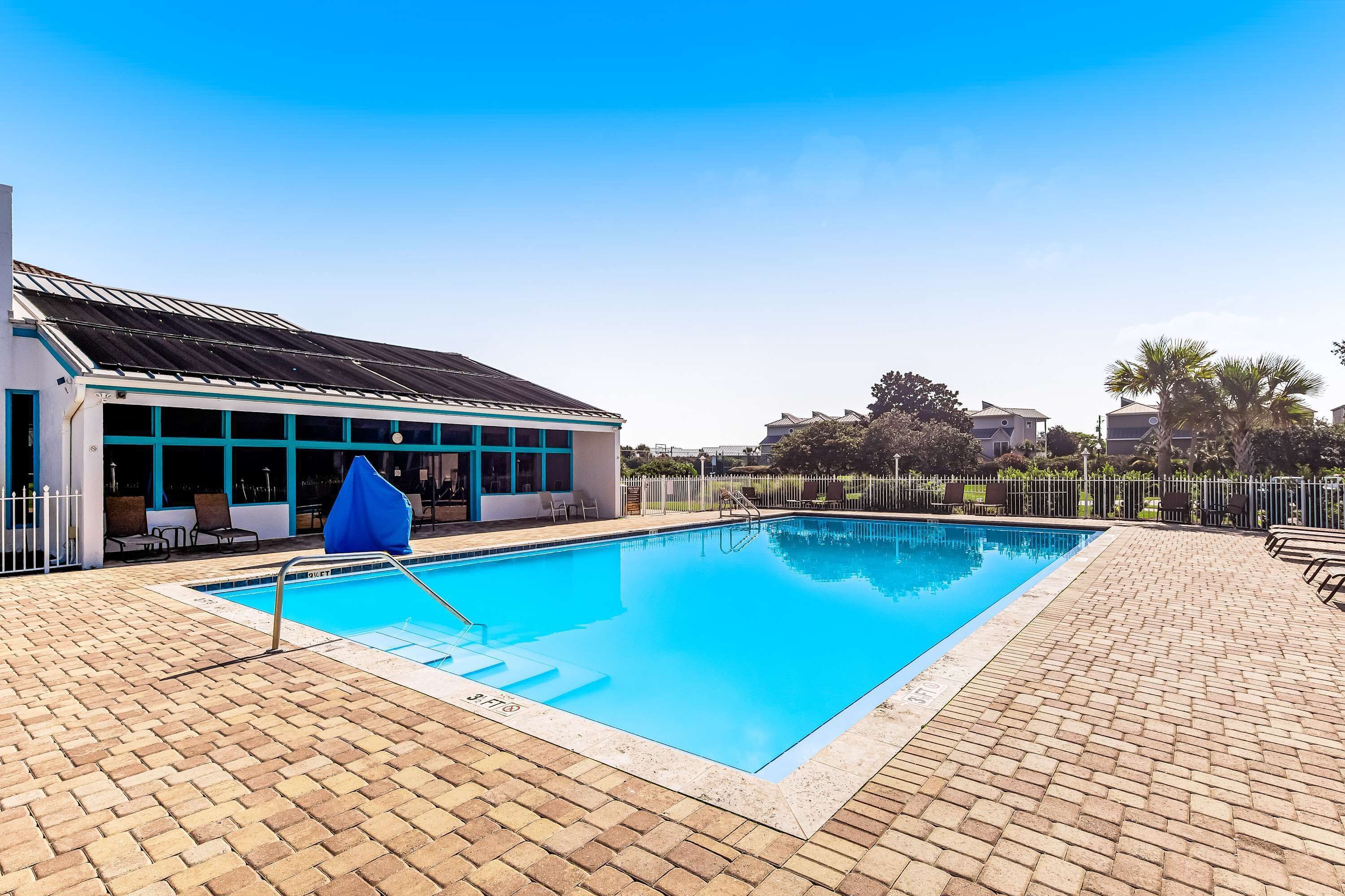 A swimming pool with a brick deck, a covered seating area, and a building with large windows in the background.