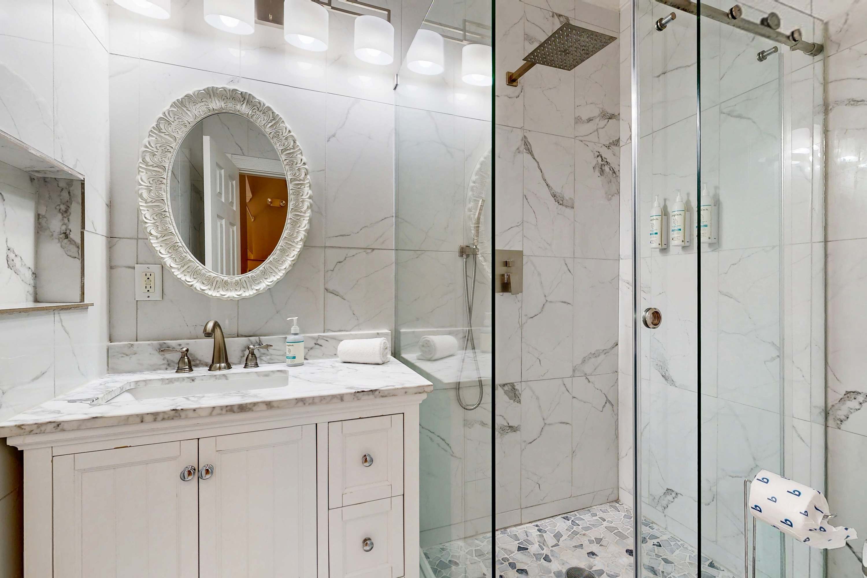 Un baño con lavamanos de mármol, un espejo grande y ornamentado, y zona de ducha con mampara de cristal.