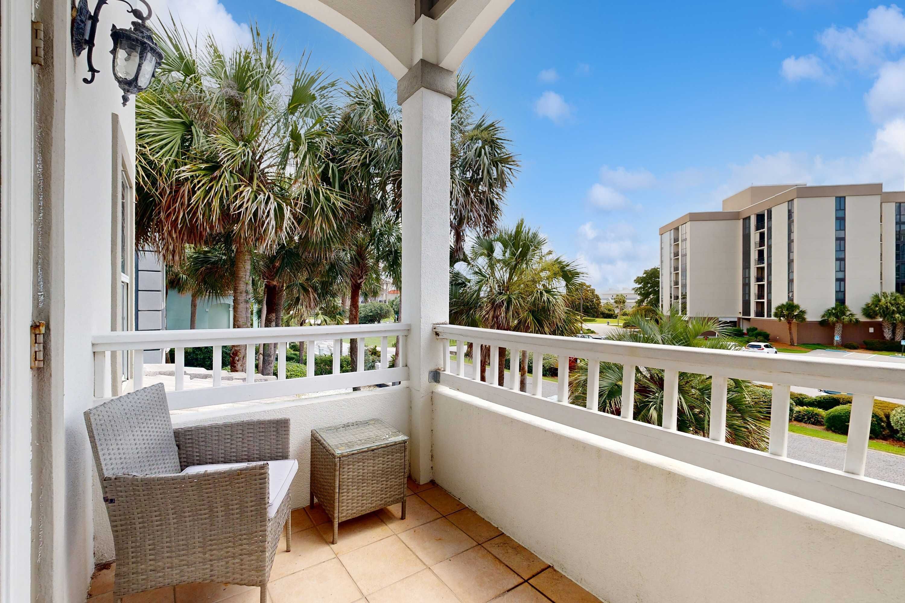 A balcony with wicker furniture, a white railing, and a view of palm trees and a building.