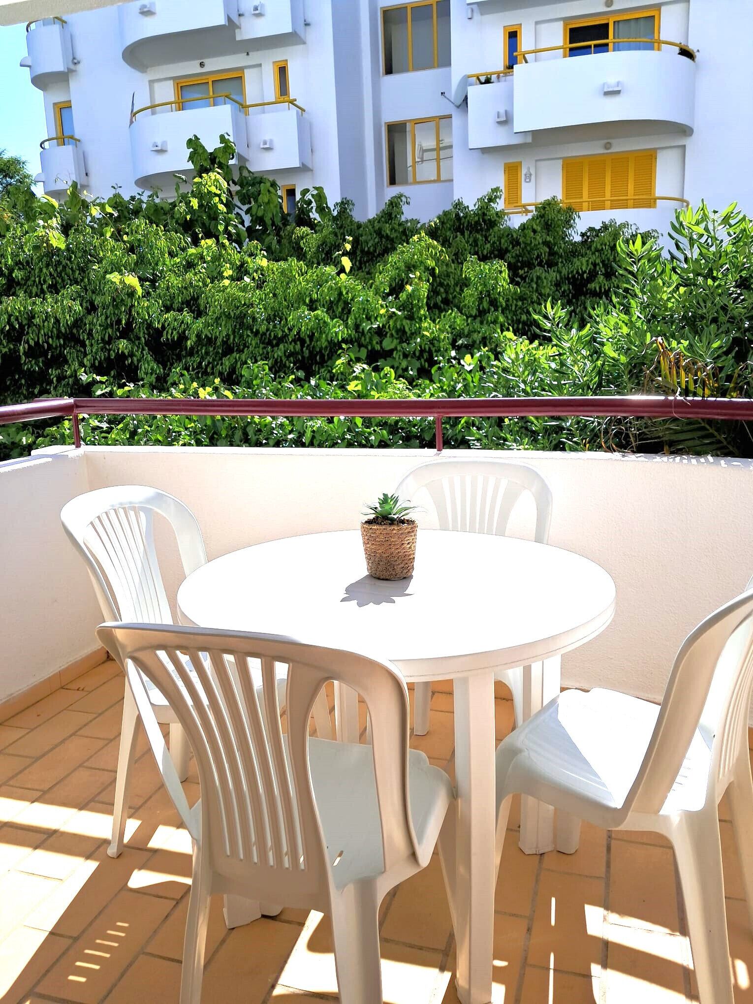 A round table with white plastic chairs and a potted plant on a balcony with a view of a white building and green foliage.