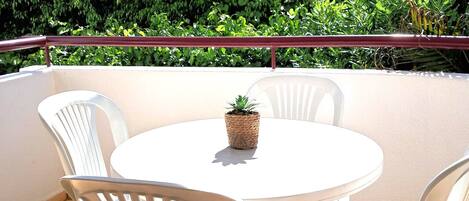 A round table with white plastic chairs and a potted plant on a balcony with a view of a white building and green foliage.